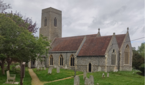 Horstead church and its church yard shown with an overcast sky and a large tree, and a path through he grass with graves either side