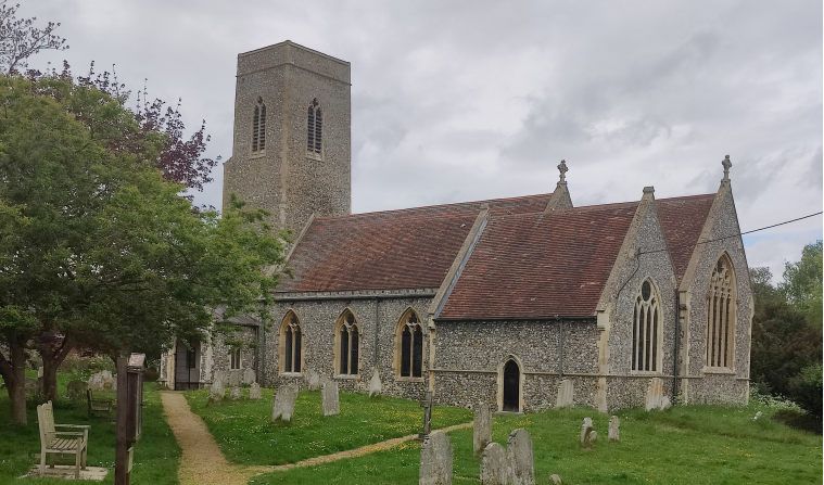 Horstead church and its church yard shown with an overcast sky and a large tree, and a path through he grass with graves either side
