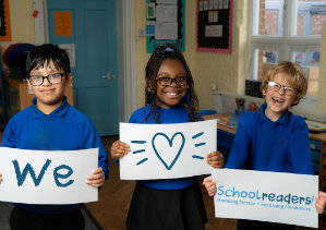 three primary age children in uniform smiling and holding up cards that say we love reading
